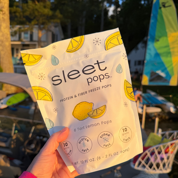 A hand holds a bag of Sleet Pops Tart Lemon Pops in an outdoor setting with a sailboat and trees in the background.