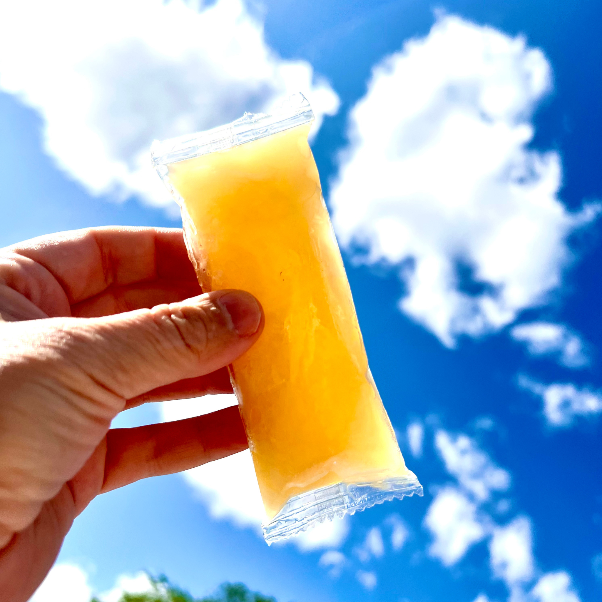 Hand holding a yellow Sleet Pop against a blue sky with clouds