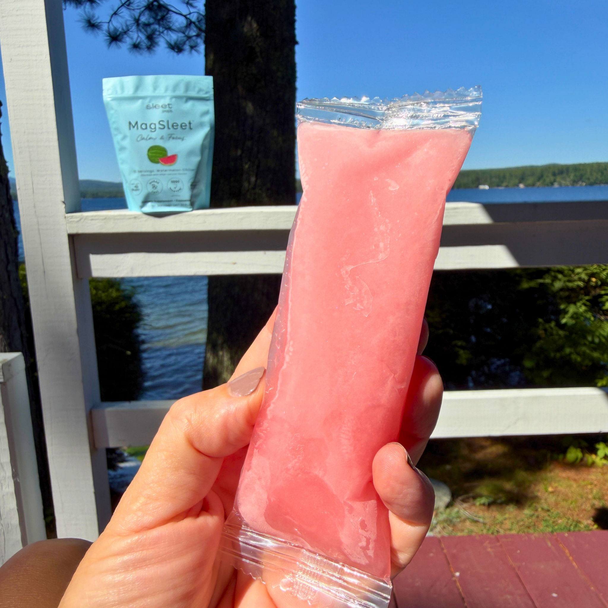 A hand holds a pink watermelon MagSleet freeze pop pack outdoors near a lake, with a branded light blue pouch resting on a white railing in the background. Made with magtein, saffron, and l-theanine. 